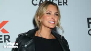 Woman with blonde hair wearing a black leather jacket and necklace smiling at a cancer awareness event backdrop