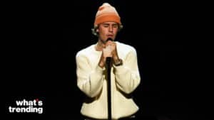 Justin Bieber singing into a microphone wearing a white sweater and orange beanie against a black background