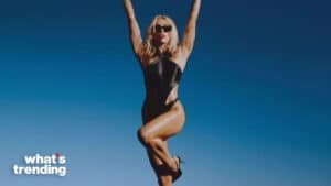 Woman in black swimsuit and sunglasses posing with arms raised against clear blue sky
