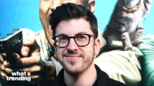 Man with glasses and beard smiling in front of a movie poster featuring a person holding a gun and a cat wearing a chain collar