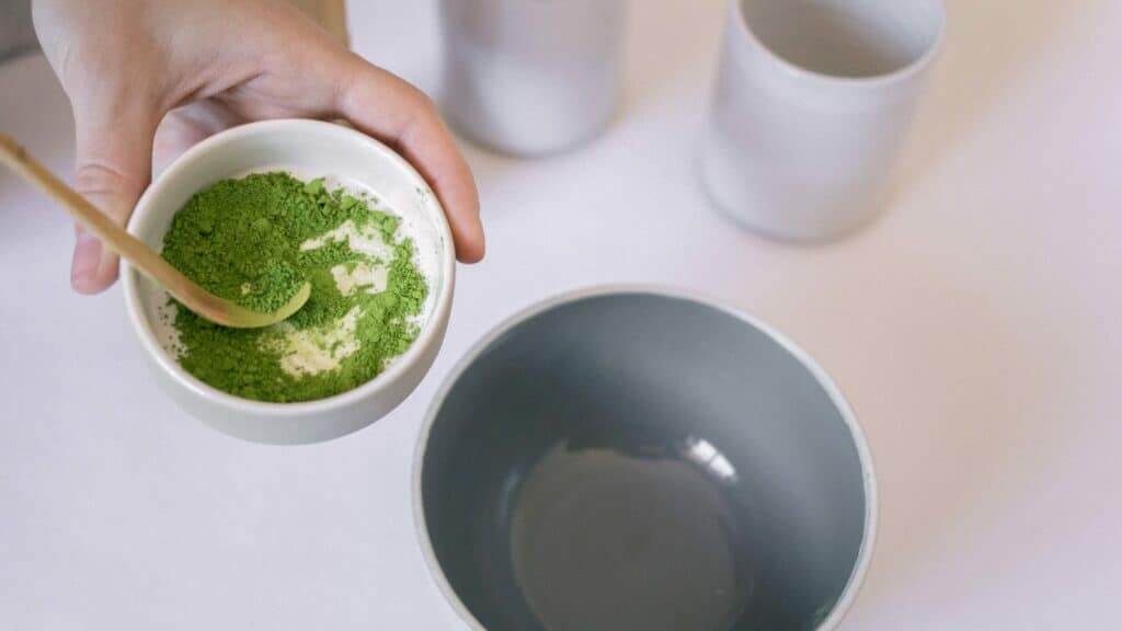 Hand holding a bowl with green matcha powder and a wooden spoon above an empty gray bowl on a white surface