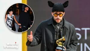 Bad Bunny wearing a black coat and hat with ears, holding a Grammy award and speaking at a podium.