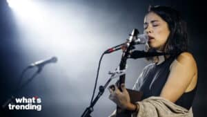 Female musician singing and playing guitar on stage with microphone and spotlight, "what's trending" logo visible