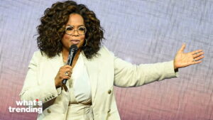 Oprah Winfrey speaking into a microphone with her arm extended, wearing a light-colored suit and glasses.