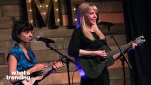 Two women performing music on stage, one playing a ukulele and the other playing a guitar and singing.