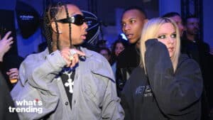 Tyga wearing sunglasses and jewelry talking to Avril Lavigne in a dark hoodie at an event with blue lighting