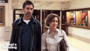 Man and woman standing indoors with concerned expressions, woman holding a brown handbag.