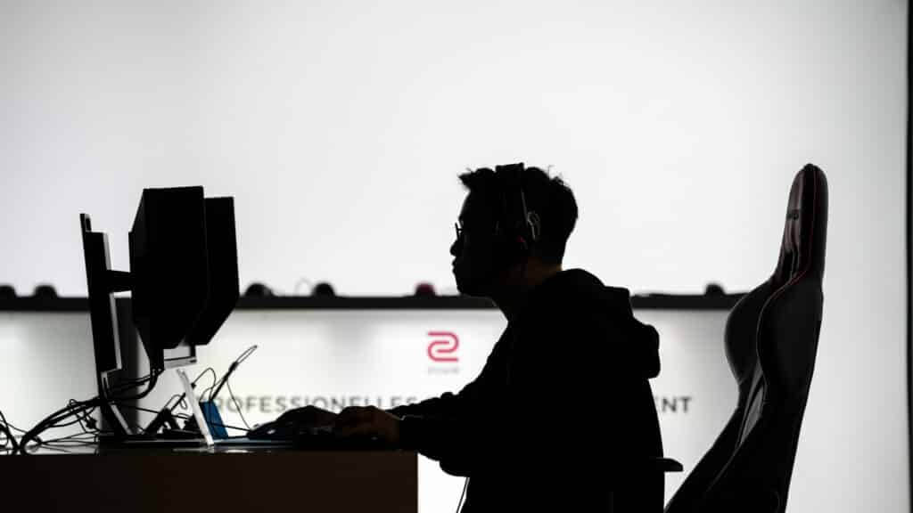 Silhouette of a person wearing headphones sitting at a desk with dual monitors and a gaming chair.