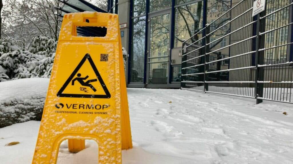 Yellow Vermop caution sign covered in snow warning of slippery surface outside a building entrance.