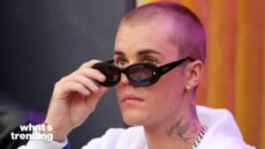 Justin Bieber adjusting black sunglasses, wearing white hoodie and gold earring, with neck tattoo visible
