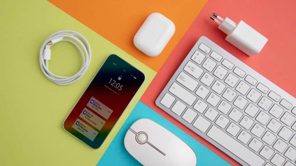 Smartphone with message notifications, white wireless earbuds case, charging cable, white keyboard, and mouse on colorful background