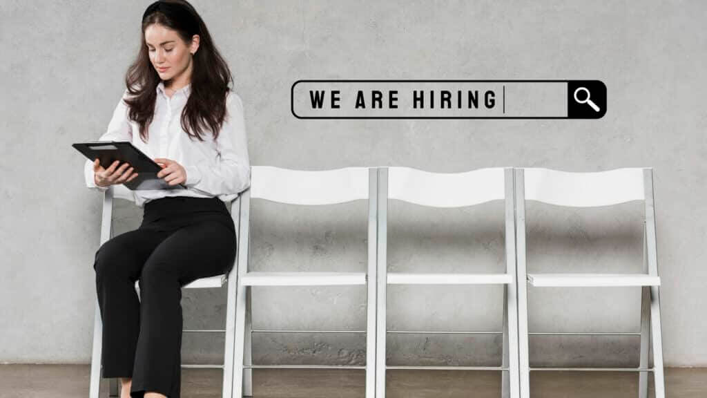 Woman in white shirt and black pants sitting on a chair holding a tablet next to a "We Are Hiring" search bar graphic.