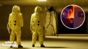 Two people in yellow hazmat suits facing a tall, thin spider-like creature in a dimly lit room.