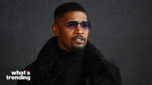Jamie Foxx wearing sunglasses and a black fur-collared coat against a dark background