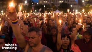Crowd holding lit candles during a nighttime vigil, one person wearing a rainbow wristband.