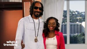 Snoop Dogg and a woman smiling together on the set of What's Trending with a cityscape background.