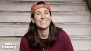 Smiling woman wearing an orange backward cap and maroon shirt sitting on stairs with "what's trending" text in corner