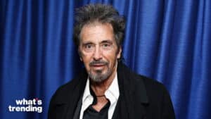 Al Pacino wearing a black coat and white shirt standing in front of a blue curtain with "what's trending" text
