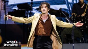 Musician performing on stage wearing a beige jacket and brown shirt with arms outstretched.