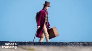 Man in a purple coat and top hat walking with a wooden suitcase against a clear blue sky