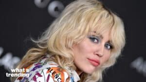 Close-up of a woman with blonde hair and blue eye makeup wearing a floral Balenciaga outfit against a dark background.