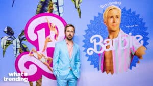 Ryan Gosling in a light blue suit standing in front of a Barbie movie promotional poster.