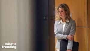 Woman in gray blazer holding a black portfolio standing against a wooden wall, looking to the side.