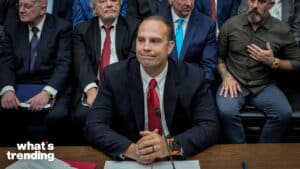 Man in a suit with a red tie sitting at a table with a microphone, surrounded by men in formal and casual attire