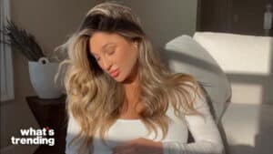 Woman with long wavy blonde hair in white top sitting on a couch with sunlight and "what's trending" text in corner