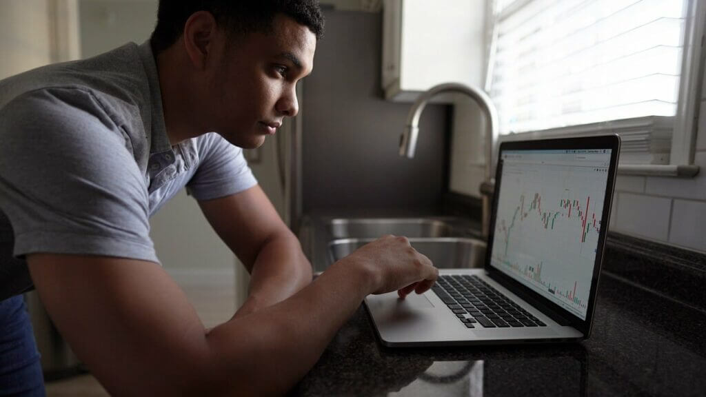 Man in gray shirt analyzing stock market charts on a laptop in a kitchen.