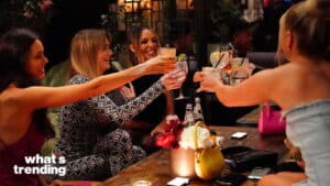 Four women toasting with cocktails at a dimly lit bar table with flowers and handbags.