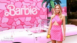Margot Robbie in a pink polka dot dress posing next to a pink vintage car with a Barbie backdrop