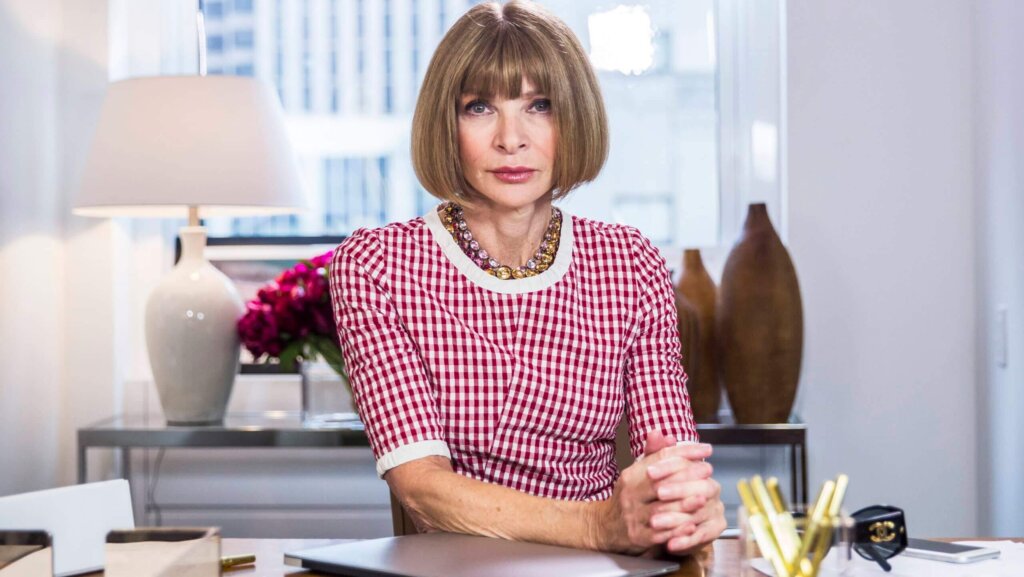 Anna Wintour sitting at a desk wearing a red and white checkered dress and a beaded necklace.