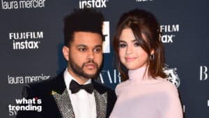 The Weeknd and Selena Gomez posing together at a formal event with branded backdrop
