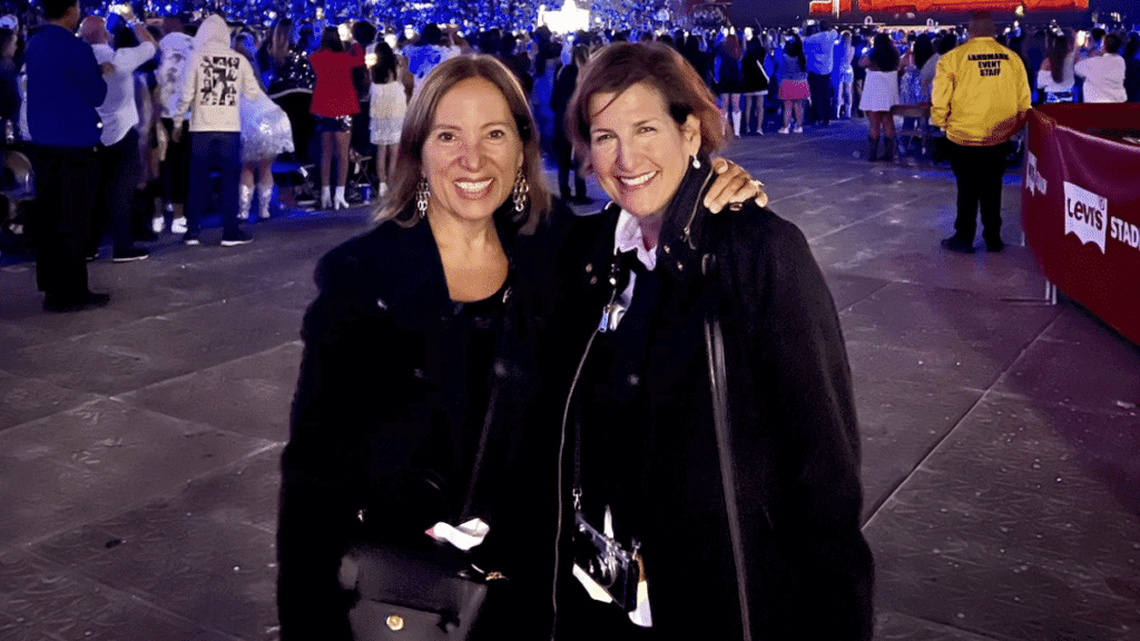 Two smiling women in black coats posing at Levi's Stadium event with crowd and staff in background