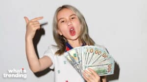 Young woman holding fan of US hundred-dollar bills making finger gun gesture with mouth open, "what's trending" logo visible