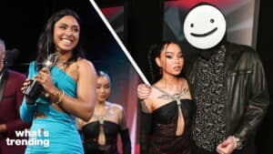 Two women in black dresses and one woman in a blue dress holding an award, with a man wearing a smiley face mask and leather jacket