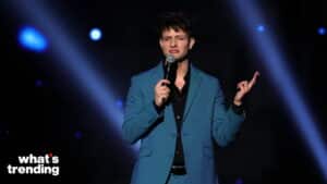Comedian holding a microphone and performing on stage with "what's trending" logo in the corner