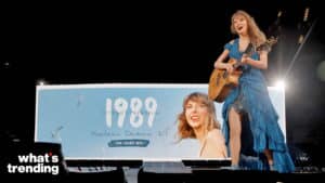 Taylor Swift performing live with guitar in front of a "1989 Taylor's Version" album promotion screen