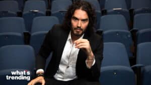 Russell Brand sitting in a theater with blue seats, wearing a black suit and white shirt.