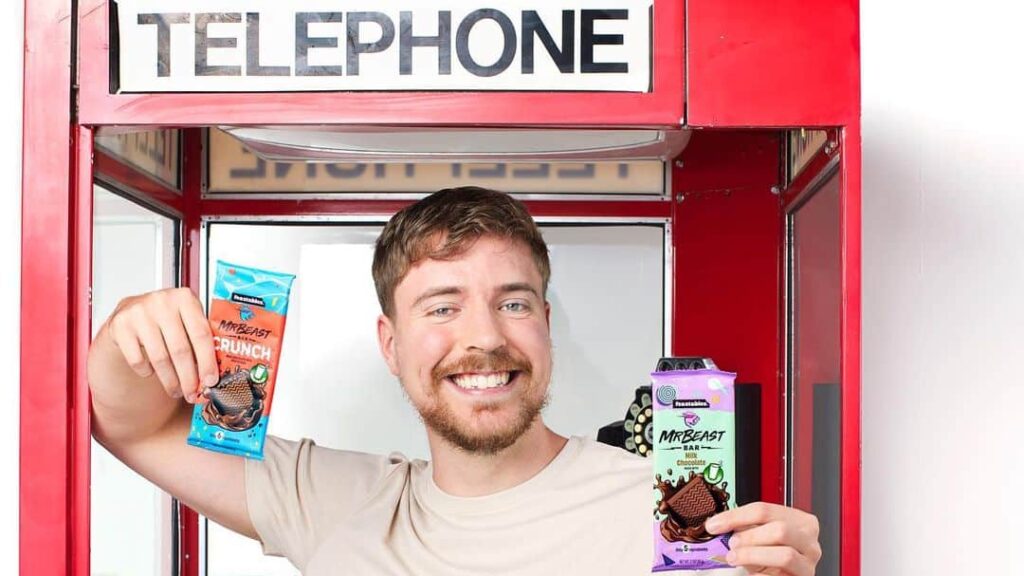 MrBeasts smiles and holds up two Feastables chocolate bars in an English telephone booth.