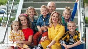 Smiling family of eight sitting on outdoor stairs, including a woman in a yellow suit and children in colorful clothing.
