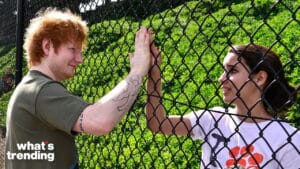 Ed Sheeran and a woman touching hands through a chain-link fence outdoors with "what's trending" logo.