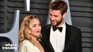 Miley Cyrus in white dress and Liam Hemsworth in black tuxedo smiling at each other at an event.