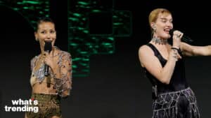 Two female performers singing on stage, one wearing a bejeweled top and the other in a black outfit with a jeweled belt.