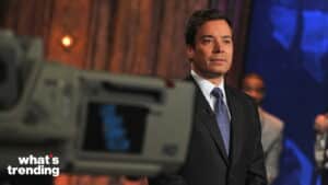 Jimmy Fallon in a suit standing on the set of the TV show "What's Trending" with a camera in the foreground.