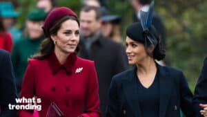Kate Middleton in a red coat and beret walking and talking with Meghan Markle in a black outfit and feathered hat