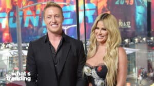 Two smiling hosts standing in front of a colorful "What's Trending" backdrop, the man in a black suit and the woman in a floral dress