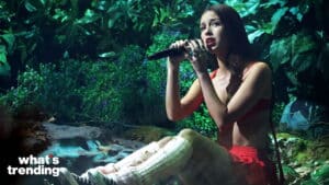 Young woman in red outfit singing into a microphone while sitting outdoors surrounded by greenery