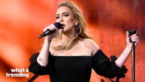 Adele singing on stage wearing a black off-shoulder dress with large hoop earrings and holding a microphone.